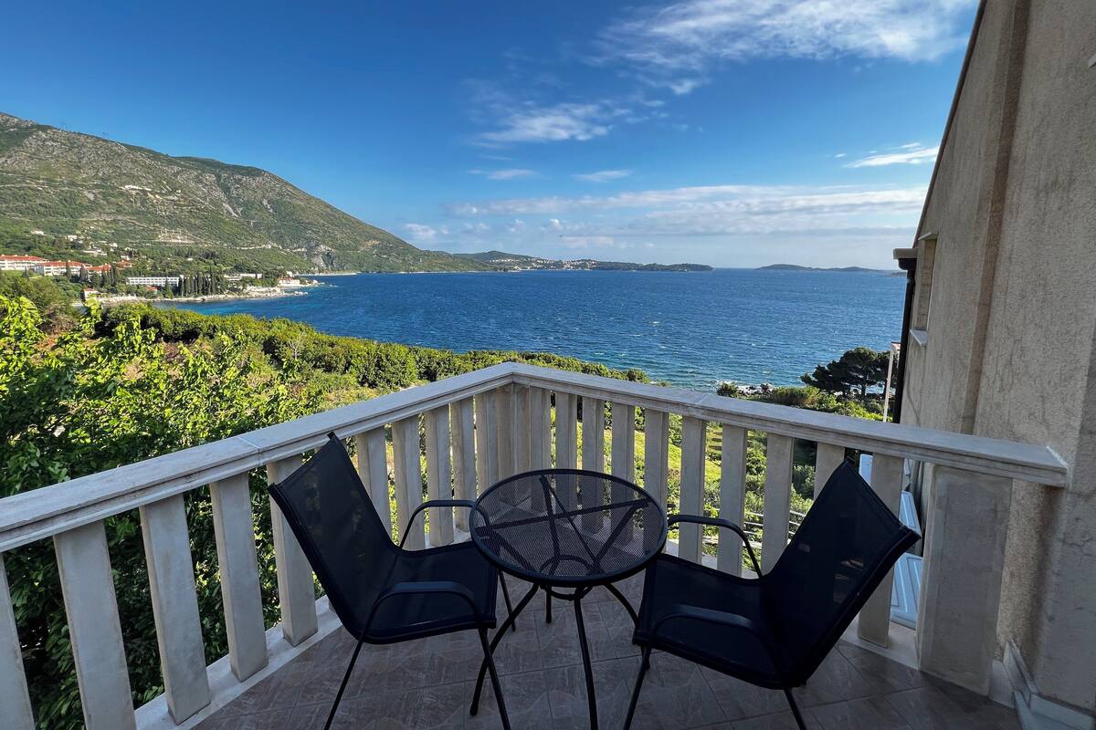 A balcony with two chairs and a table, overlooking a coastal landscape with hills and a clear blue sea.