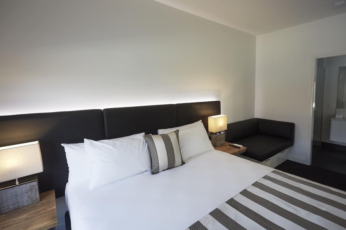 Una habitació d'hotel amb un llit gran, dues taules de nit amb làmpades, un capçal de llit fosc i una petita zona de seients amb un coixí.