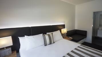 Habitación de hotel con una cama grande, dos mesitas de noche con lámparas, un cabecero oscuro y una pequeña zona de estar con un cojín.