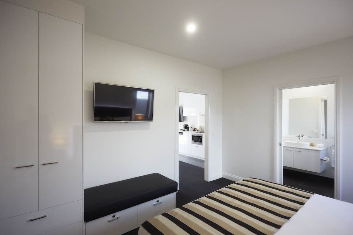 Habitació d'hotel moderna amb un llit, una televisió penjada a la paret, un bany amb mirall i una zona de cuina equipada.