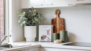 Cocina con armarios blancos, una planta en maceta, tabla de cortar de madera, libro de cocina y una taza verde.