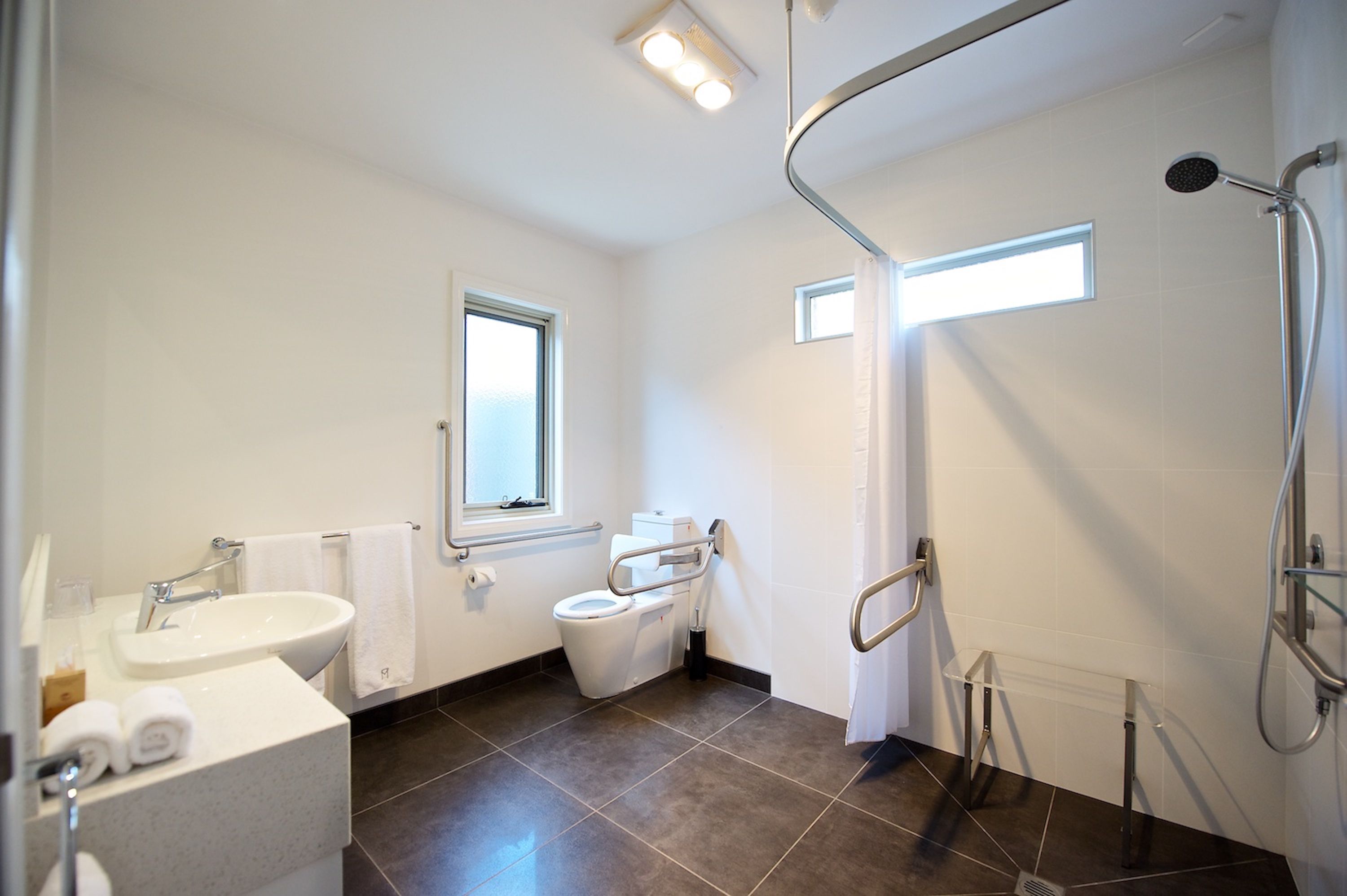 A modern bathroom with a sink, toilet, and shower. It includes a window, a towel rack, and a grab bar for safety.