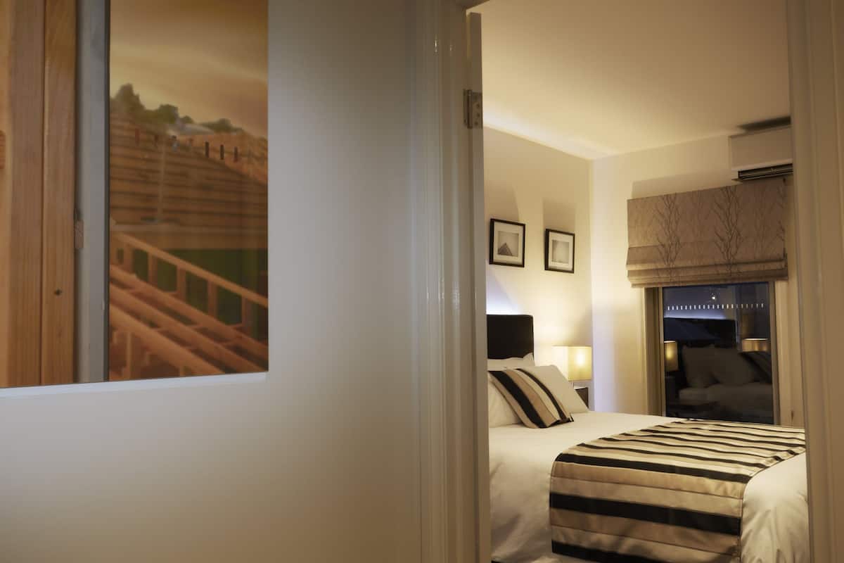 Habitació d'hotel amb un llit, una finestra que mostra una estructura de fusta, una tauleta de nit amb una làmpada i una finestra amb una persiana.
