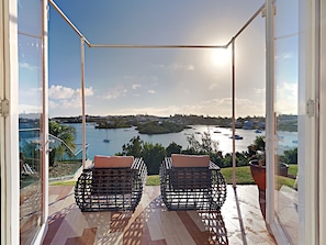Een balkon met twee rieten stoelen en een glazen deur, met uitzicht op een jachthaven met boten en een glooiend landschap.