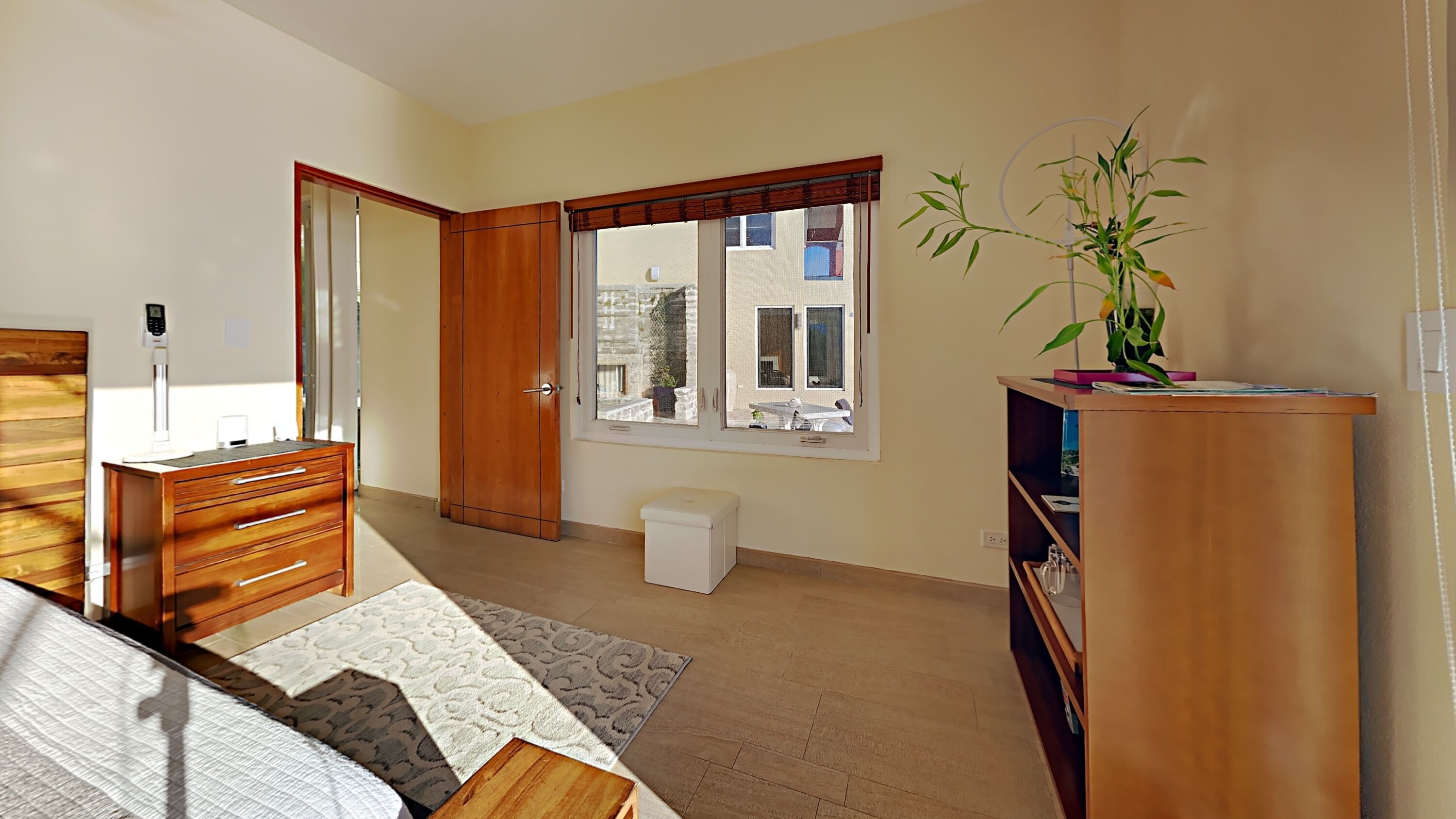 A bedroom with a wooden wardrobe, a bedside table, a small bedside lamp, a window with a view of buildings, and a potted plant.