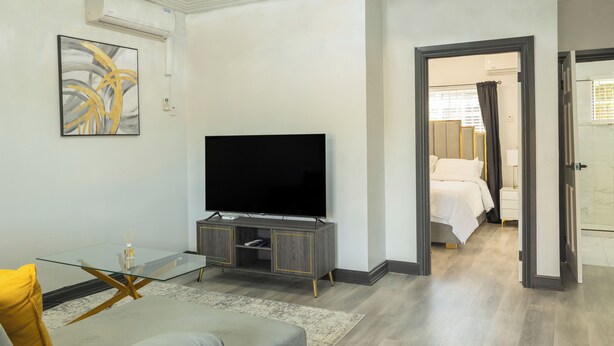 A modern living room with a flat-screen TV on a glass table, a sofa, and a bed in the adjacent room. - Furnished 1 bedroom Apartment in Gated Complex  (Kingston)