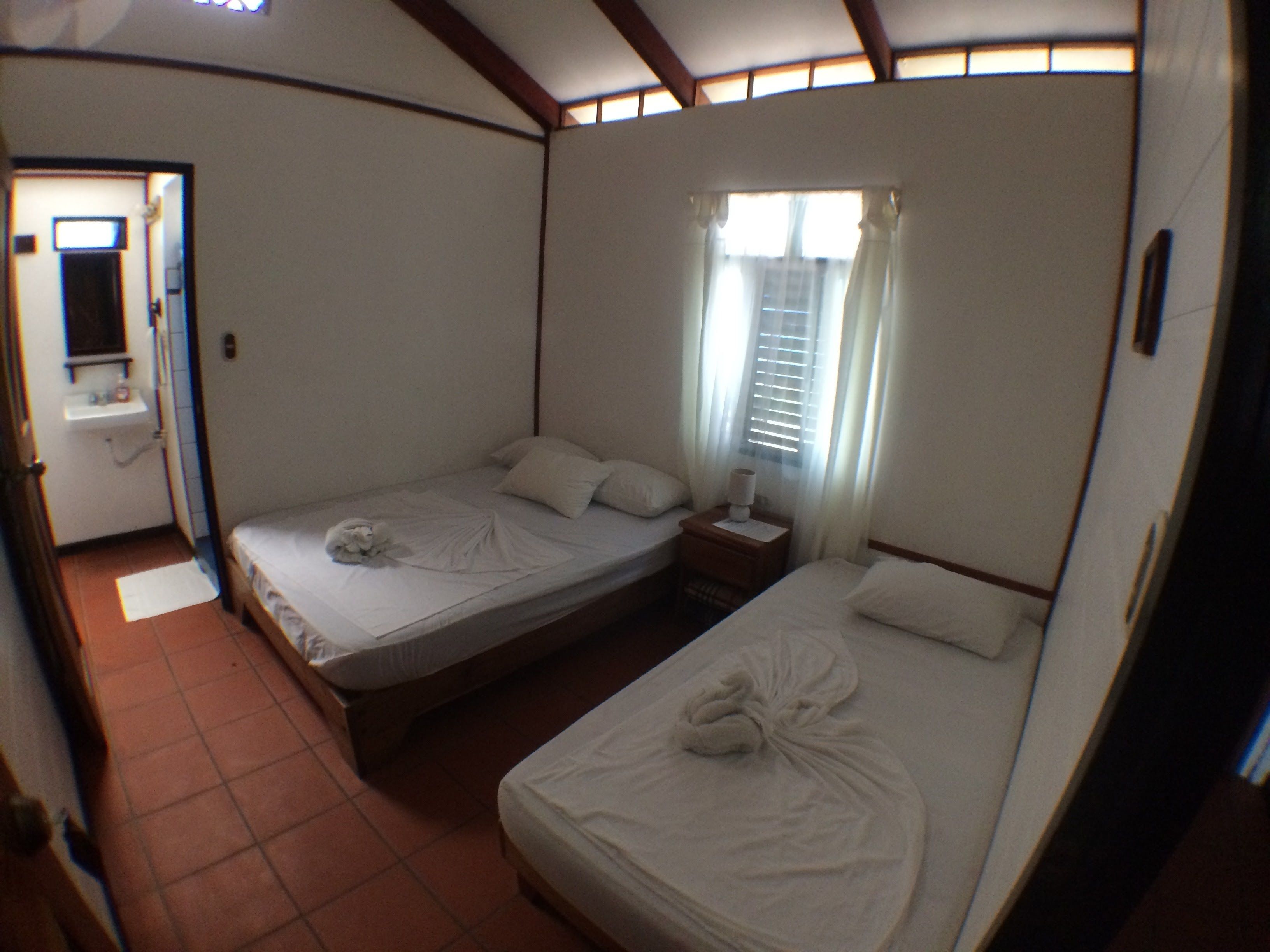 Habitación de hotel con dos camas, una mesita de noche con lámpara, ventana con cortinas y baño con lavamanos y espejo.