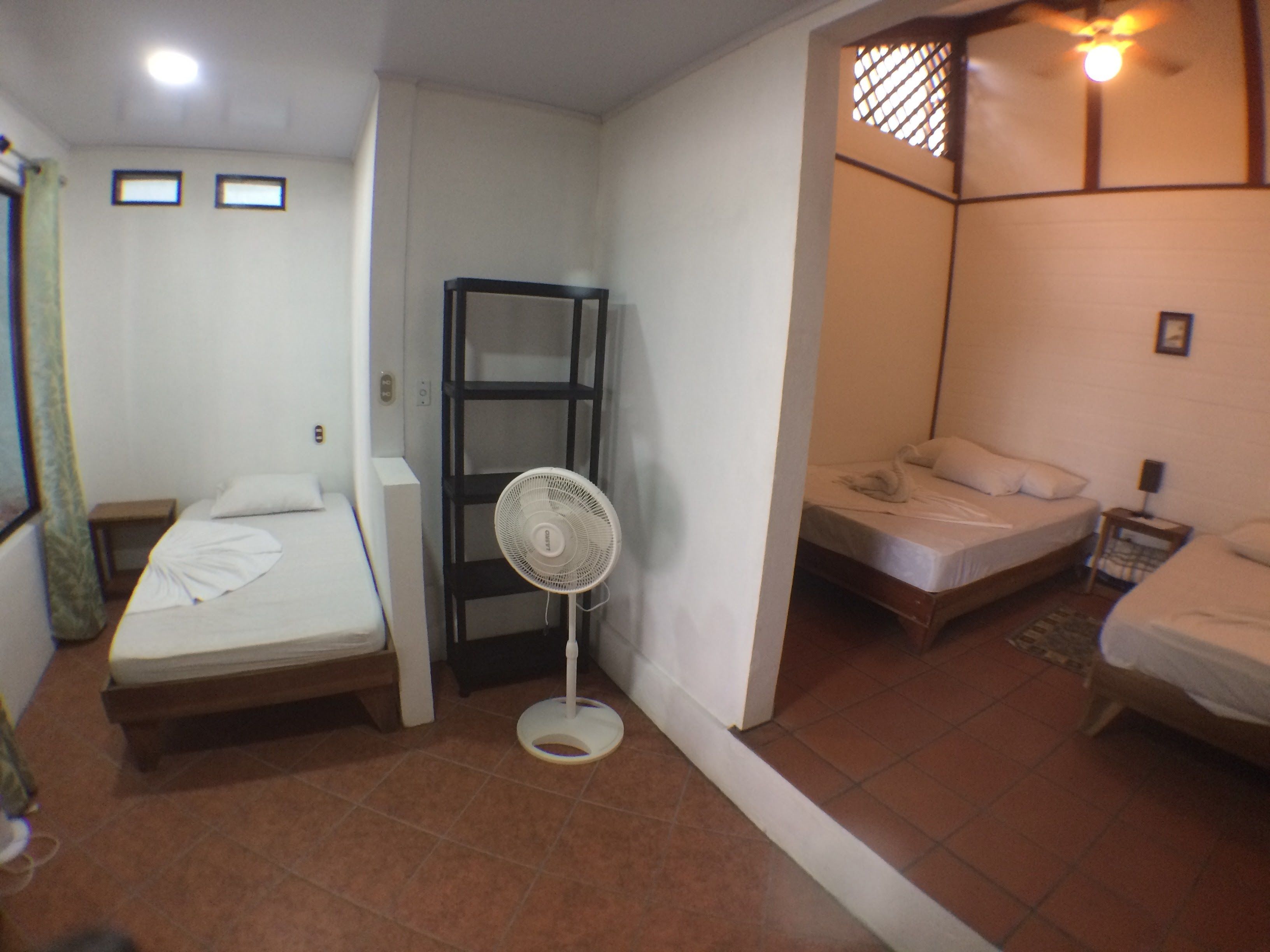 A small, compact room with two single beds, a standing fan, and a shelf unit.