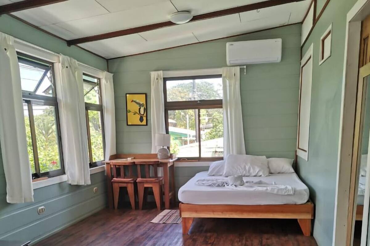 A room with a bed, a desk, and a chair. There is a window with curtains, a picture on the wall, and an air conditioning unit.