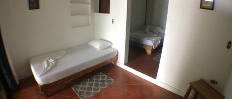 Una habitación pequeña con cama individual, suelo de baldosas, una mesita de noche y un taburete.