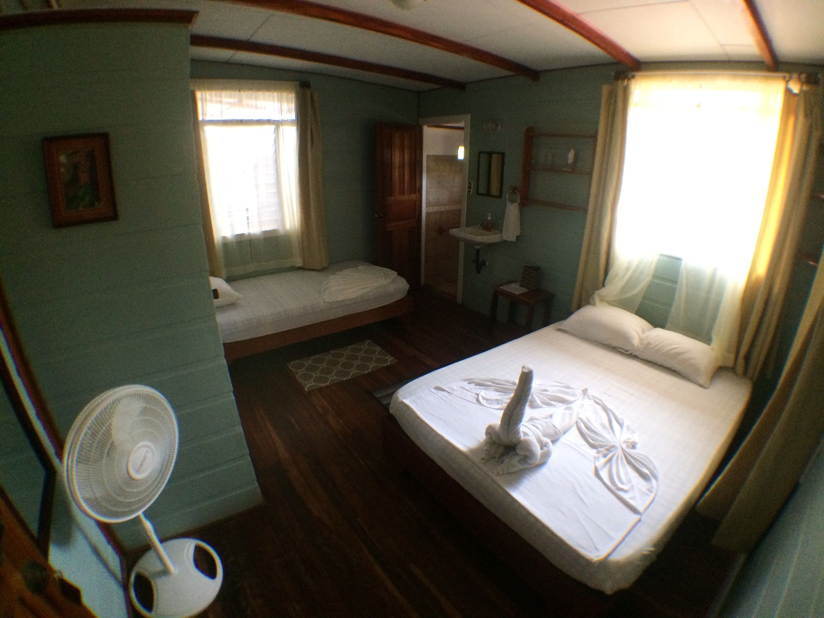 A compact hotel room with a single bed, a small sitting area, a fan, and a bathroom with a sink and mirror.