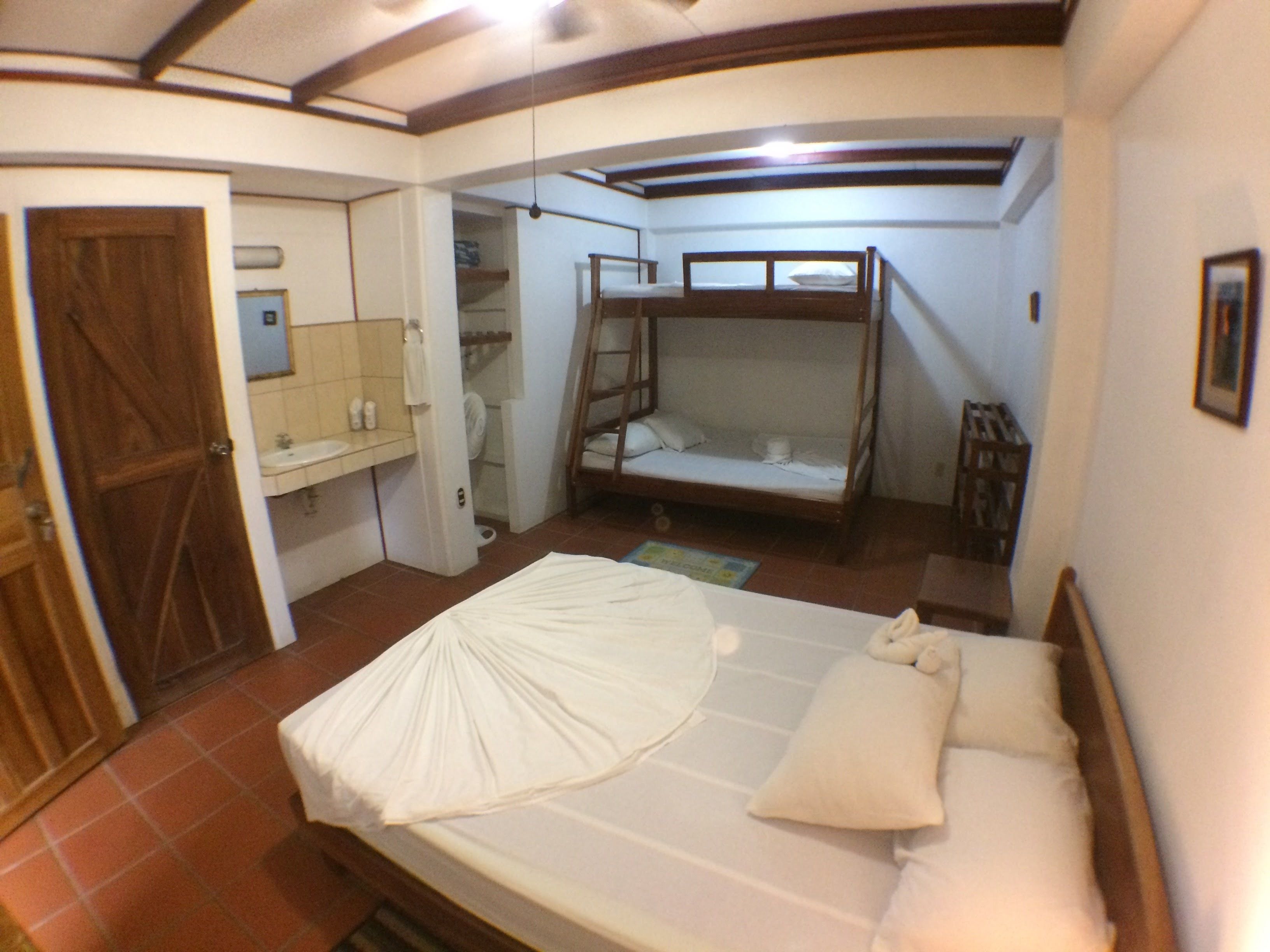 Un dormitorio con una cama litera, una puerta de madera, un baño con lavamanos y espejo, y piso de baldosas.