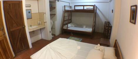 Un dormitorio con una cama litera, una puerta de madera, un baño con lavamanos y espejo, y piso de baldosas.