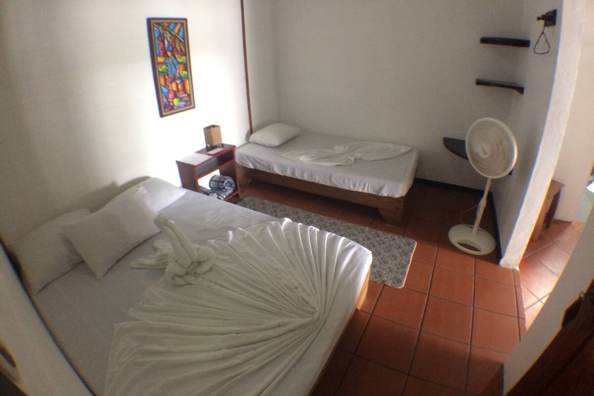 A room with a large bed, a smaller bed, a bedside table with a lamp, a fan, and a colorful painting on the wall.