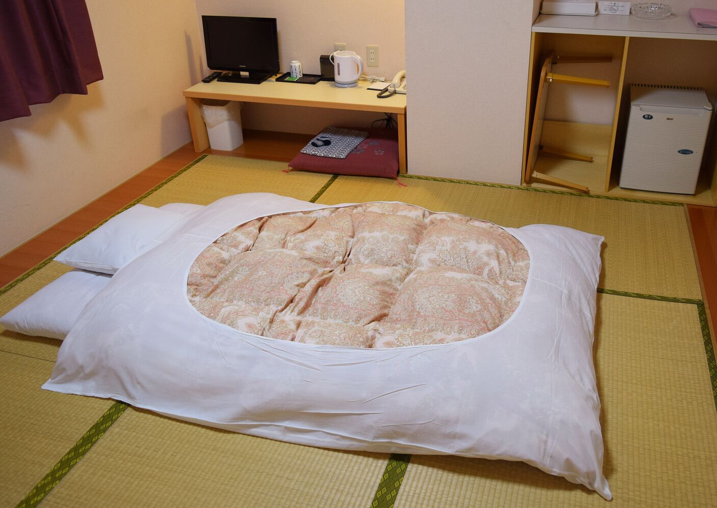 A tatami mat room with a futon bed, a desk with a computer, and a small refrigerator.
