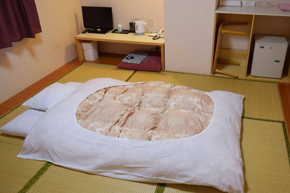 A tatami mat room with a futon bed, a desk with a computer, and a small refrigerator.