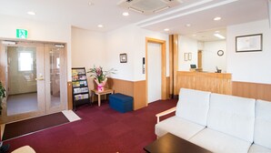 A reception area with a white couch, a wooden reception desk, a blue bench, and a bulletin board with pamphlets. - All In Stay Hakodate (Hakodate)