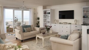 A living room with a sofa set, a coffee table, and a television. There is a bookshelf and a window with a view of the beach. - Bella Vista is a Stunning Beachfront Apartment with 200 5 star reviews (East Preston)