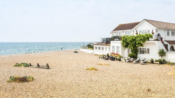 A beachfront property with a large white building, a patio with lounge chairs, and a view of the sea. - Bella Vista is a Stunning Beachfront Apartment with 200 5 star reviews (East Preston)