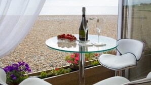 A glass table with a bottle of wine, a glass, and a bowl of strawberries on a balcony overlooking a pebble beach. - Bella Vista is a Stunning Beachfront Apartment with 200 5 star reviews (East Preston)