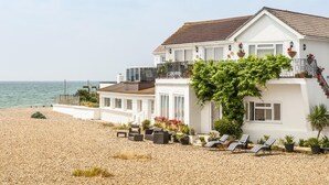 A white two-story house with a balcony, a garden with potted plants, and a pebble beach in the foreground. - Bella Vista is a Stunning Beachfront Apartment with 200 5 star reviews (East Preston)