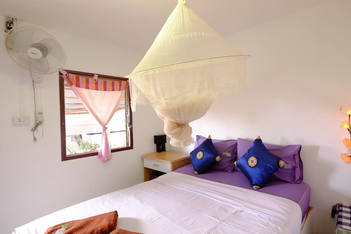 A bedroom with a bed, a bedside table, a fan, a window with curtains, and a decorative light fixture.