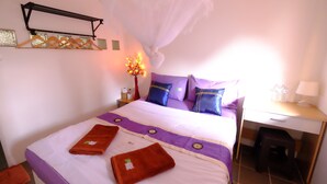 A neatly made bed with purple and white bedding, a bedside table with a lamp, and a small desk with a chair. - Patty's Secret Garden (Ko Lanta)