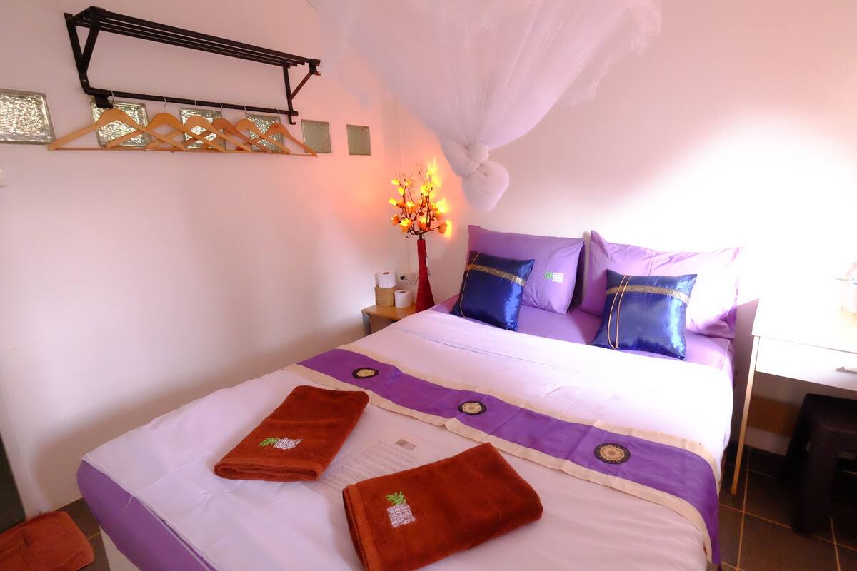 A neatly made bed with purple and white bedding, a bedside table with a lamp, and a small desk with a chair.