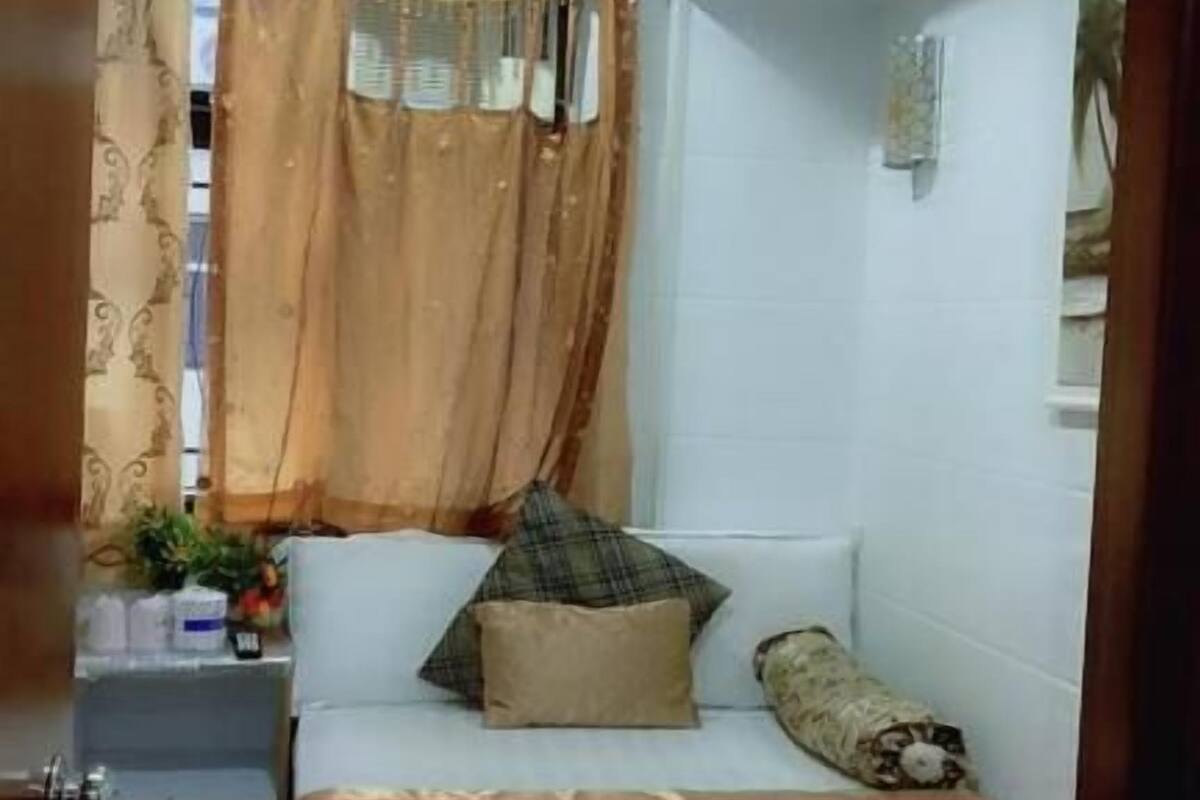 A small, compact room with a bed, a nightstand, a window with curtains, and a wall-mounted shelf.