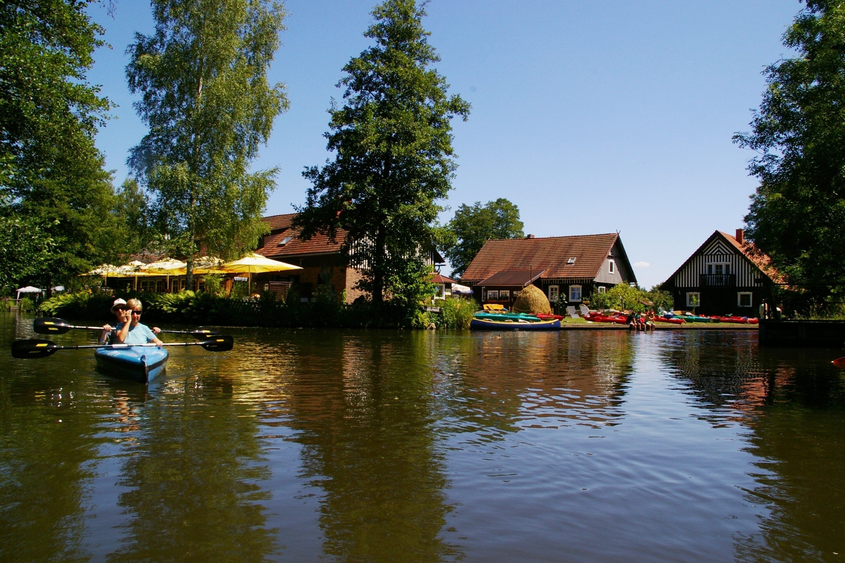Une rivière paisible où l’on peut naviguer en barque à rames ou en canoë, entourée de bâtiments traditionnels et d’une végétation luxuriante.