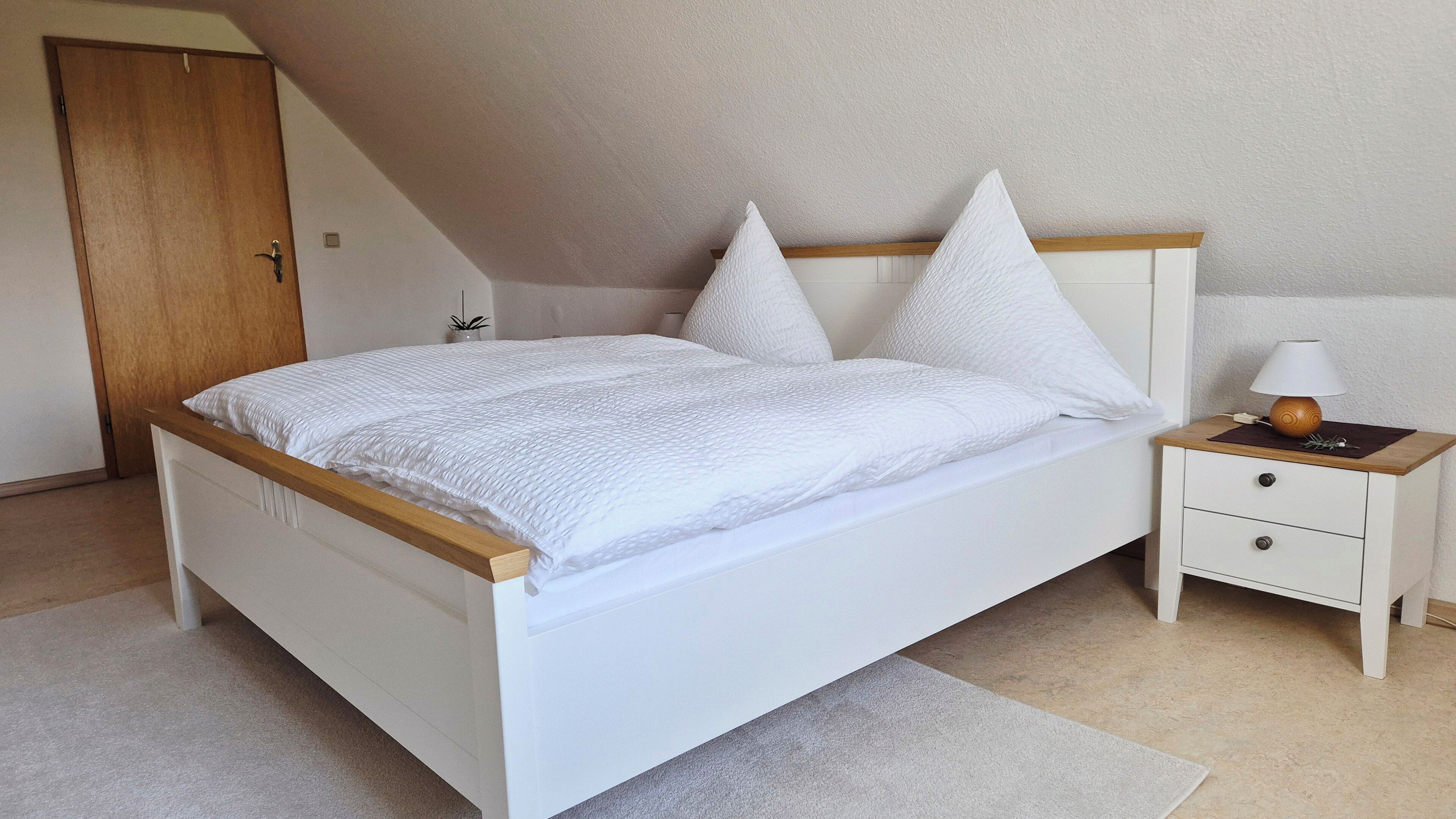 Ein Schlafzimmer mit einem weißen Bett, einem hölzernen Kopfteil, einem Nachttisch mit Lampe und einer kleinen Schublade.