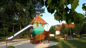 Un parque infantil con tobogán y estructuras para escalar, rodeado de árboles y un edificio de techo rojo.