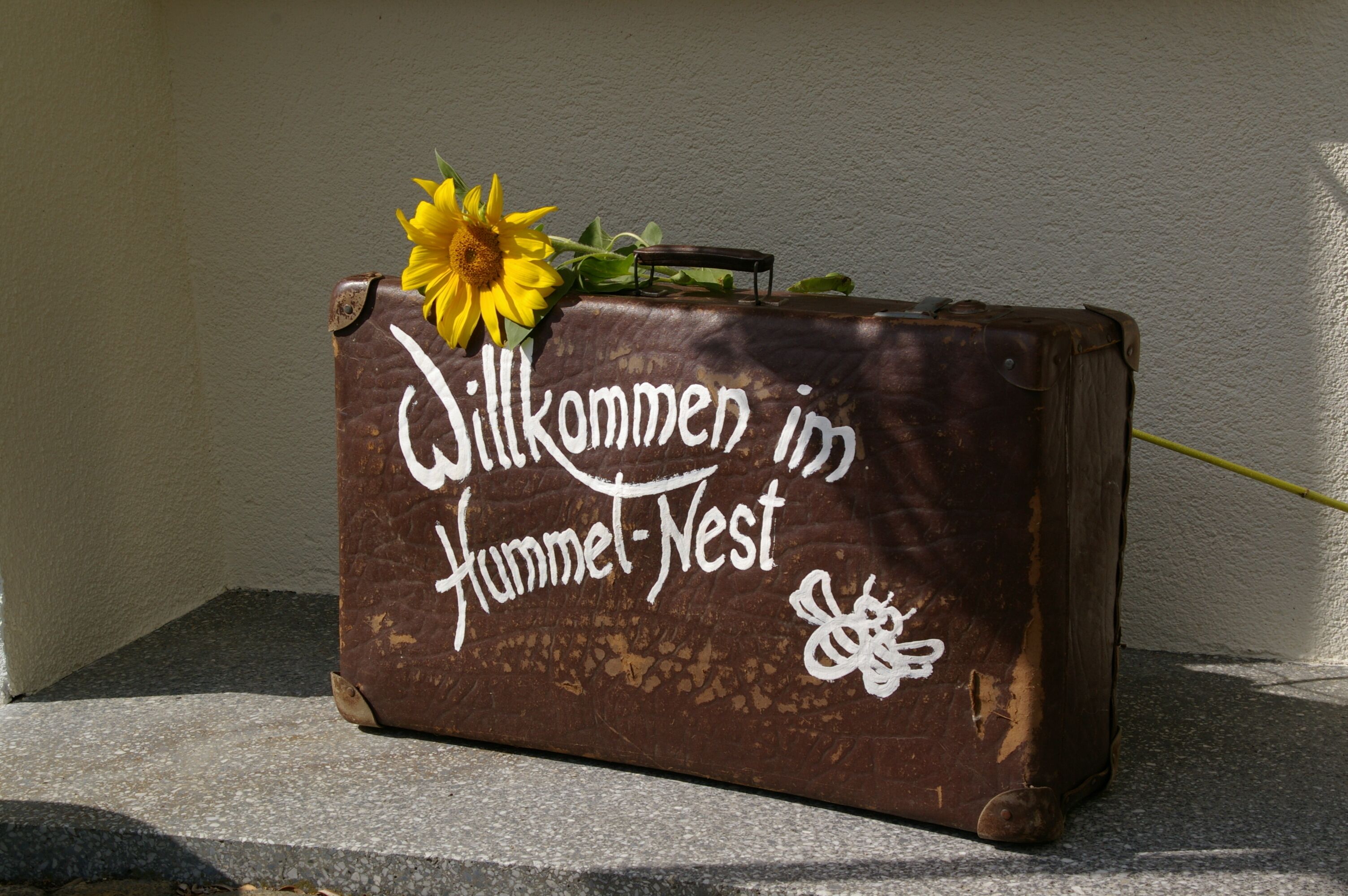 Ein Koffer mit dem Aufdruck "Willkommen im Hammel-Nest" und einer Sonnenblume darauf.
