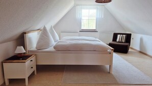 Un dormitorio con cama, mesita de noche, lámpara, sillón y ventana con vista a áreas verdes.