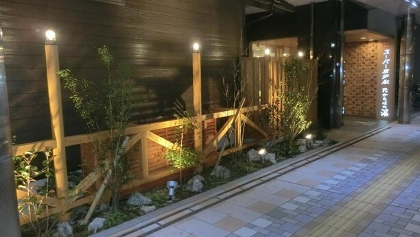 A modern building with a wooden facade, a small garden area with plants and rocks, and a tiled walkway. - Super Hotel Miyazaki (Miyazaki)