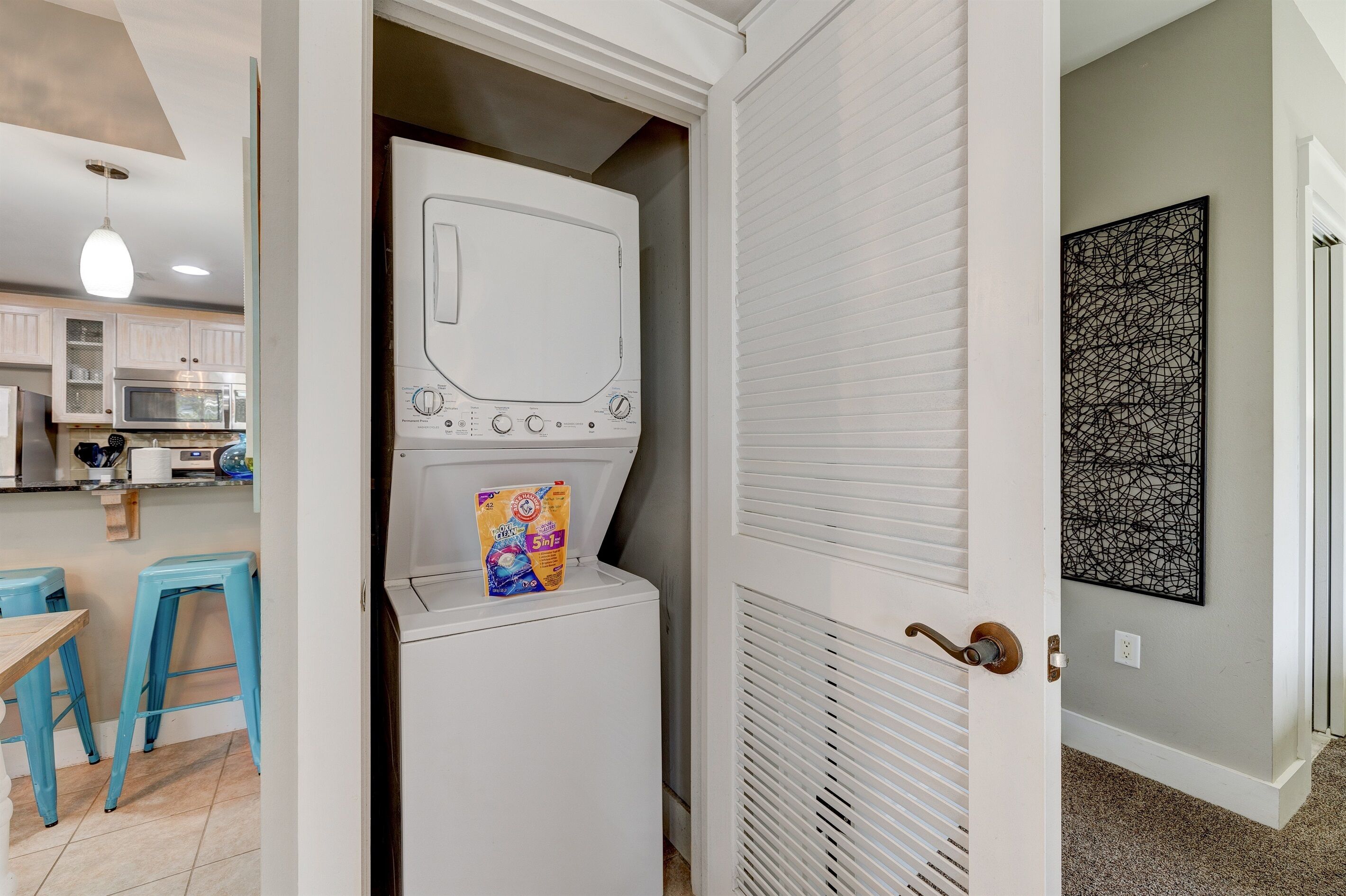 A laundry room with a washing machine, a dryer, and a door leading to a kitchen area with stools and a microwave.