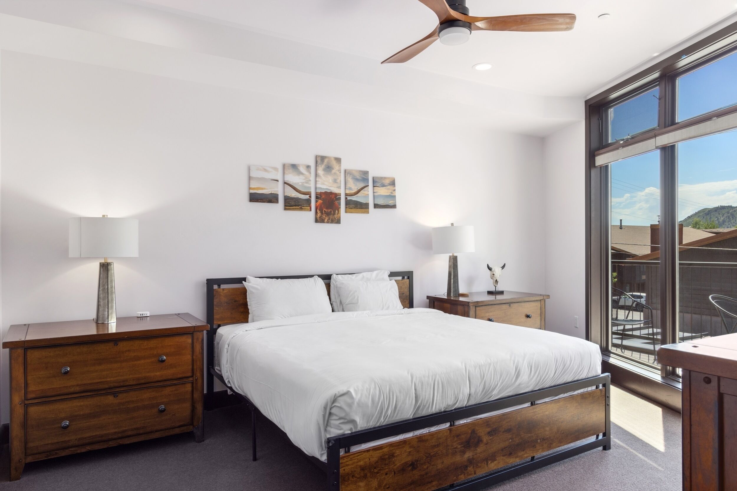 Un dormitorio con una cama, mesitas de noche de madera, un ventilador de techo y un ventanal que ofrece vistas al exterior.