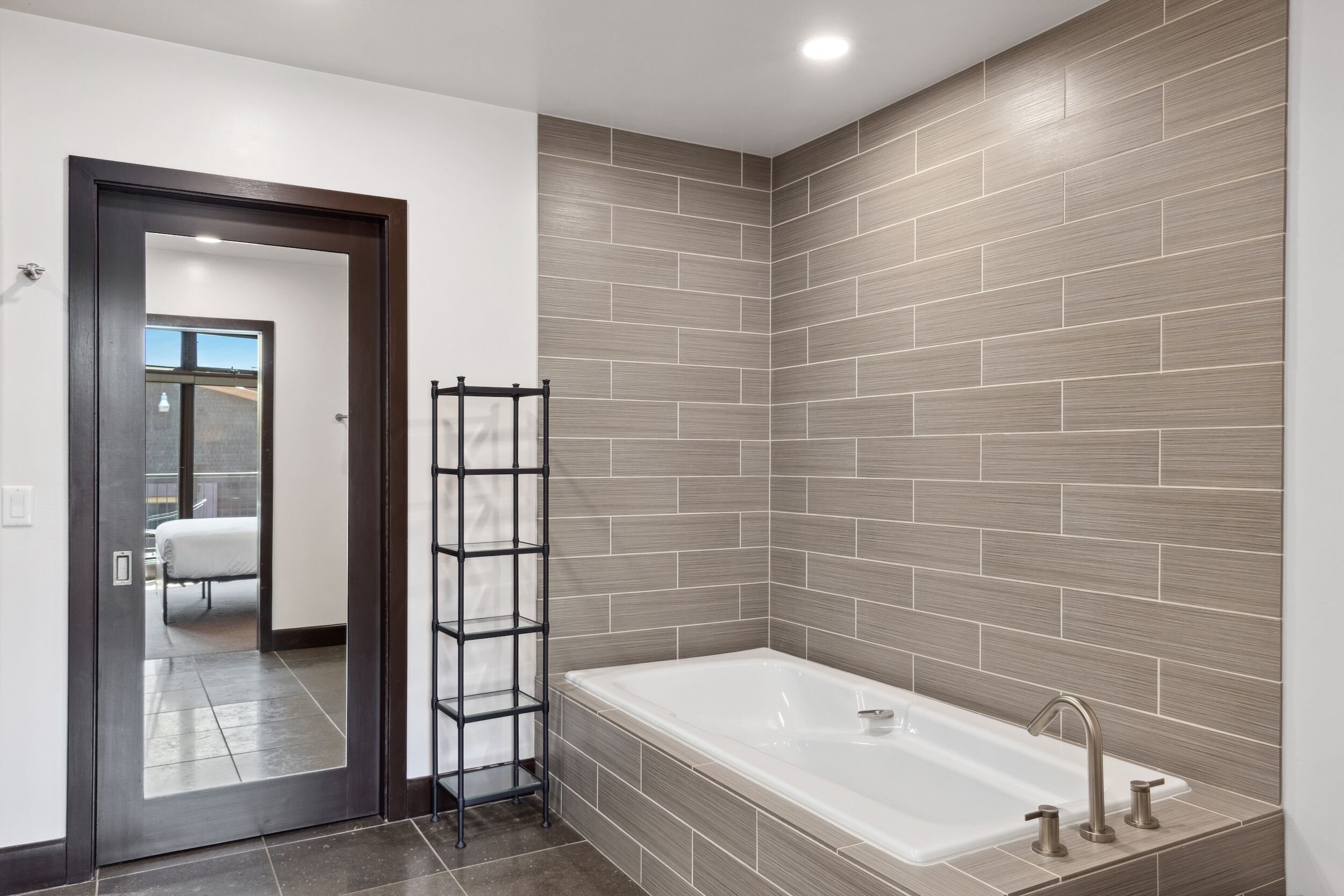 A modern bathroom with a large bathtub, a wooden shelf, and a door leading to another room.