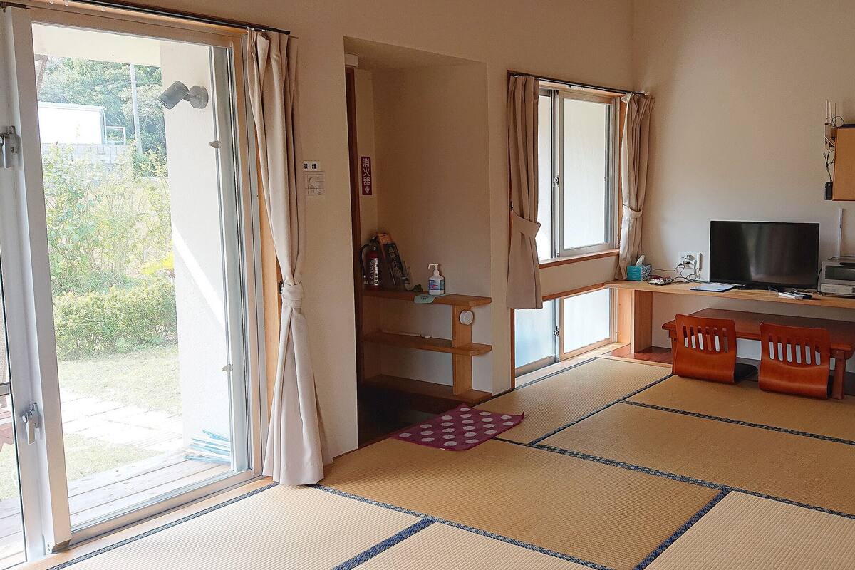 Quarto compacto com porta de vidro deslizante para uma varanda, uma pequena mesa com cadeira, televisão e frigorífico.