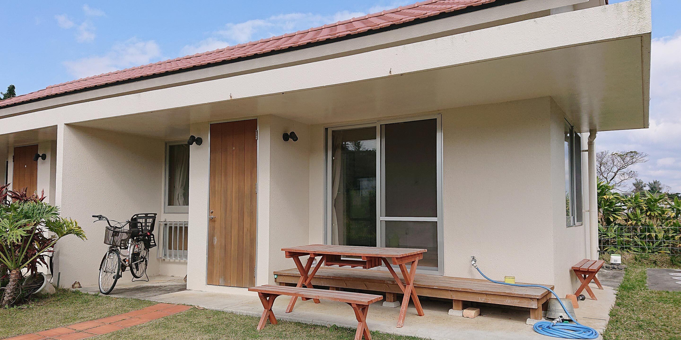 A house with a patio area featuring a wooden table and benches, a bicycle, and a garden with plants.