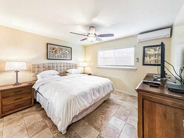 Une chambre à coucher comprenant un lit, des tables de chevet, un ventilateur de plafond, une fenêtre avec des stores et des œuvres d’art accrochées aux murs.