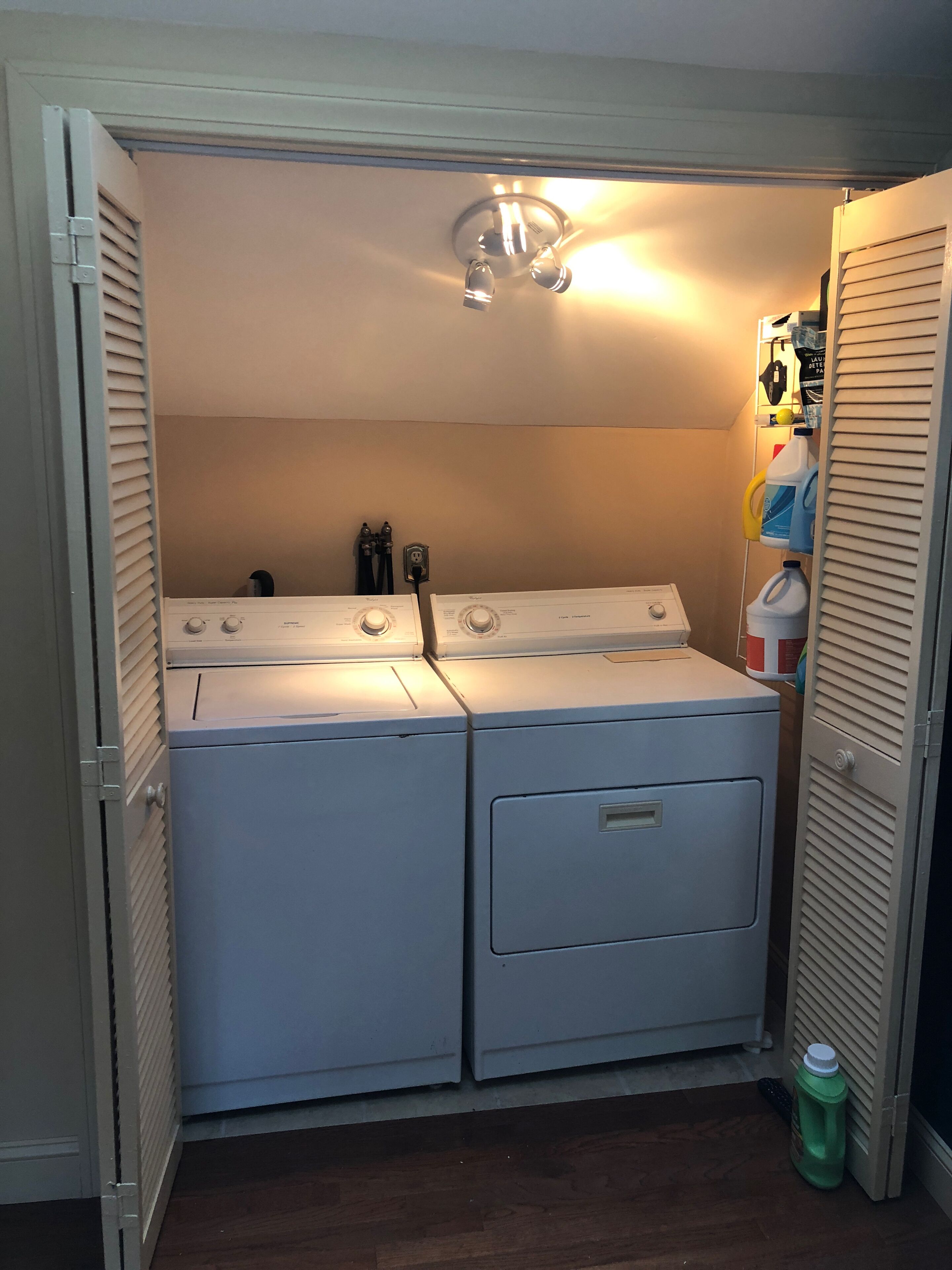 A laundry room with two stacked front-loading washers and dryers, a ceiling light, and a shelf with cleaning supplies.