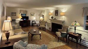 A living room with a sofa, armchairs, a coffee table, and a fireplace. There are lamps, a television, and a patterned rug on the floor.
