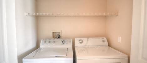 A laundry room with two white front-loading washers and dryers, a small shelf above the dryers, and a door on the left.