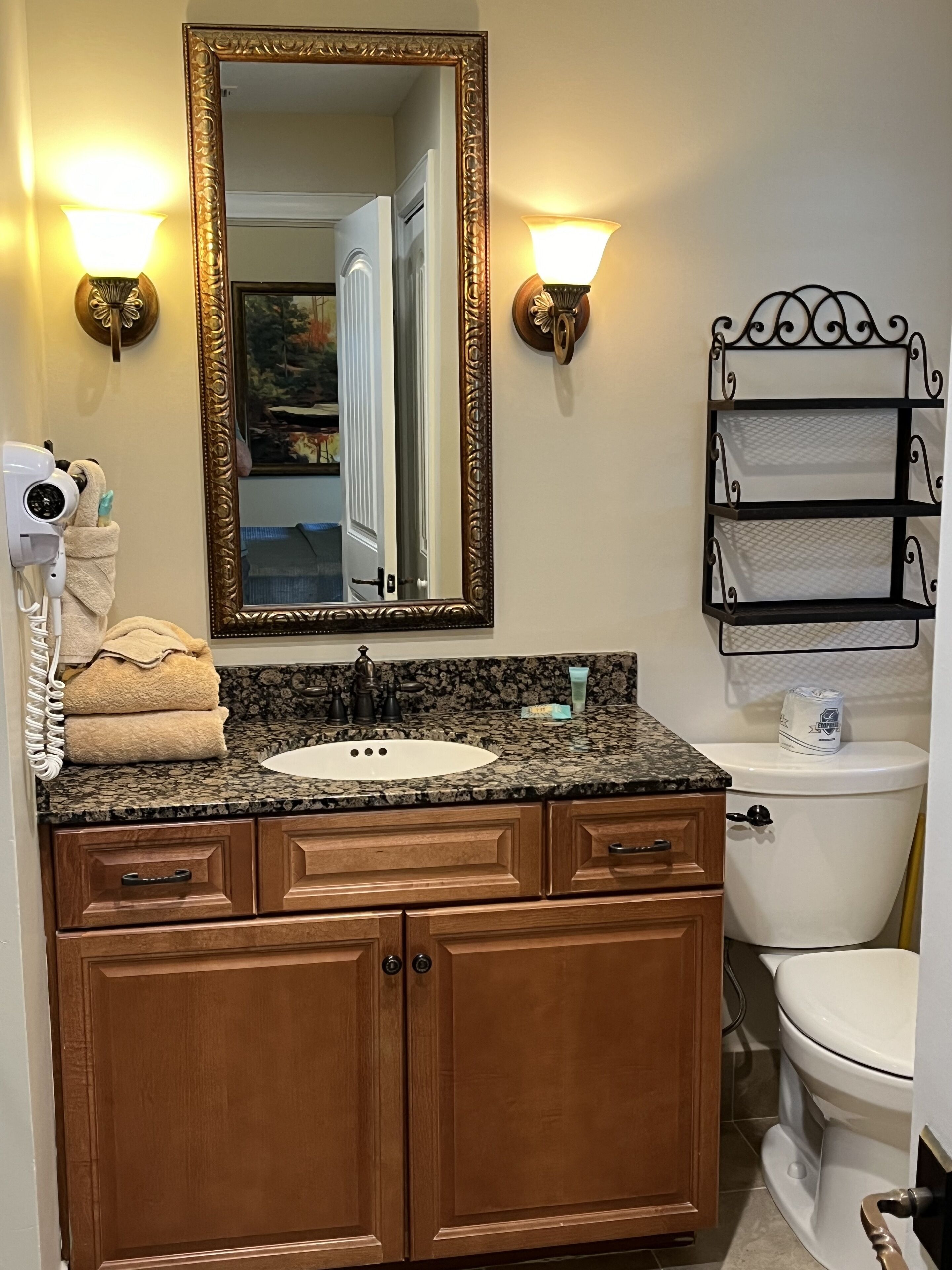A bathroom with a granite countertop sink, wooden vanity, a large mirror with a gold frame, two wall-mounted light fixtures, a towel rack, and a toilet with a metal shelf above it.