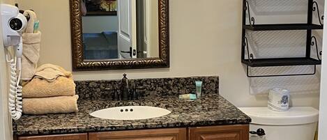 A bathroom with a granite countertop sink, wooden vanity, a large mirror with a gold frame, two wall-mounted light fixtures, a towel rack, and a toilet with a metal shelf above it.