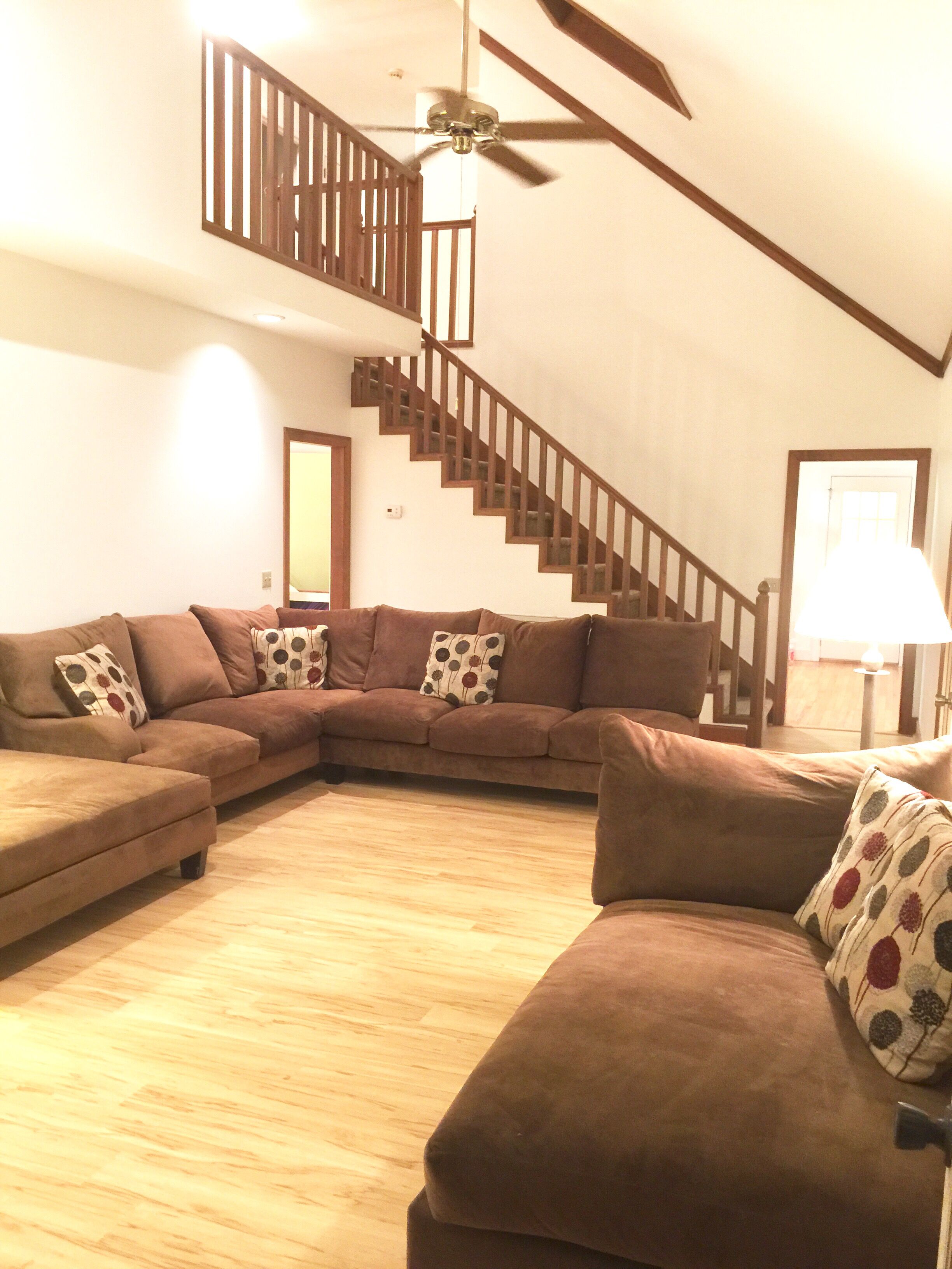 A living room with a brown sectional sofa, a wooden staircase, and a ceiling fan.