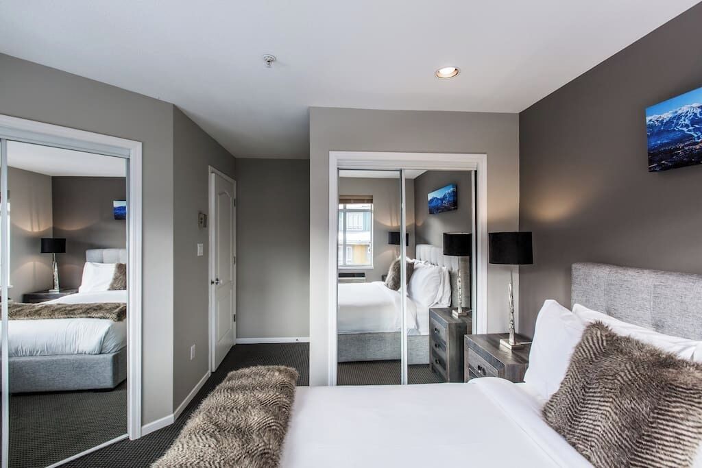 A hotel room with a bed, a desk, a chair, a mirror, and a view of another room.