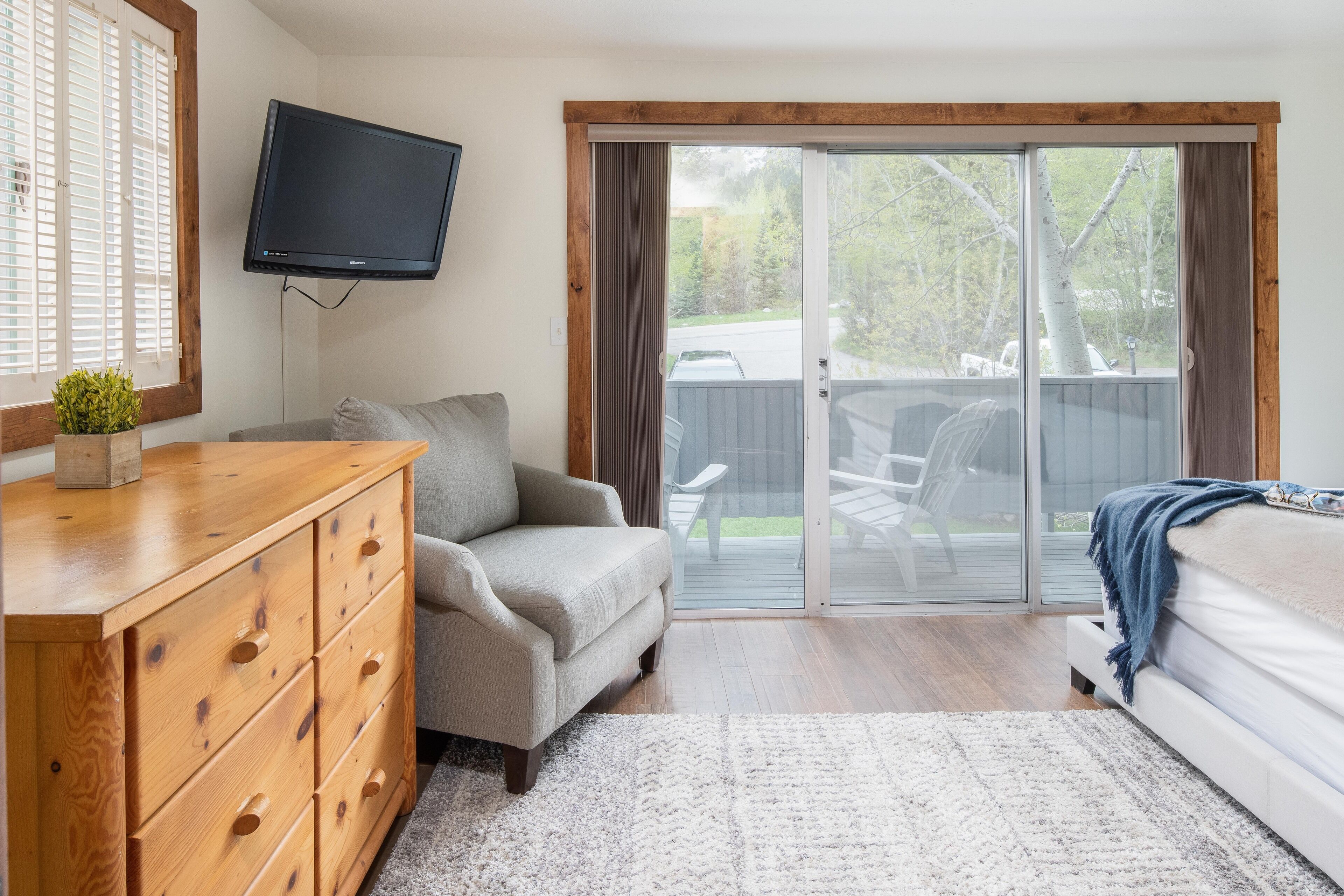 A bedroom with a wooden dresser, a gray sofa, a bed with a blue blanket, a TV mounted on the wall, and a sliding glass door leading to a backyard.