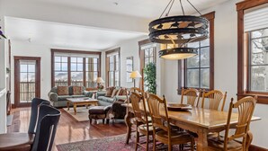 A spacious living area with a wooden dining table, chairs, and a large chandelier. There are multiple windows providing natural light and a view of the outdoors.